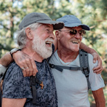 Two smiling men with arms around one another in a wooded area