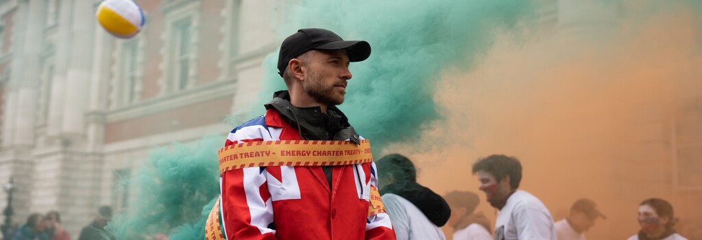 A demonstrator stands with tape reading 'Energy Charter Treaty' wrapped around them, taping them to a barrel emitting green gas