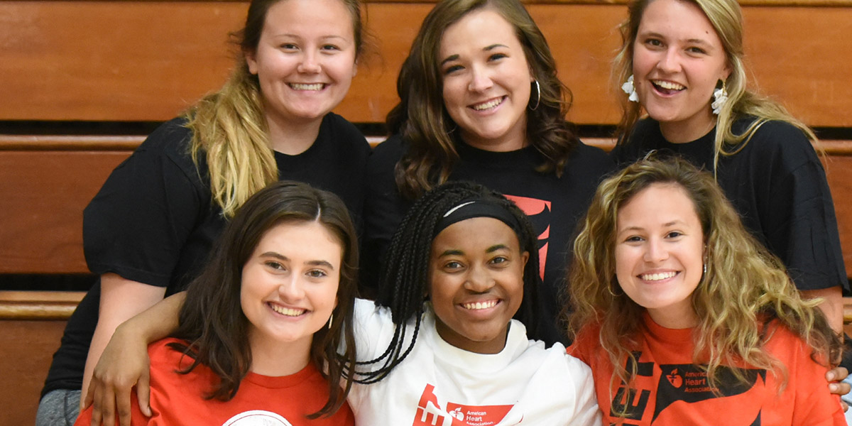 Six high school age girls sitting on stands inside a gym, three of them on one row and the other three on the row in front of and below them, smile toward the camera.